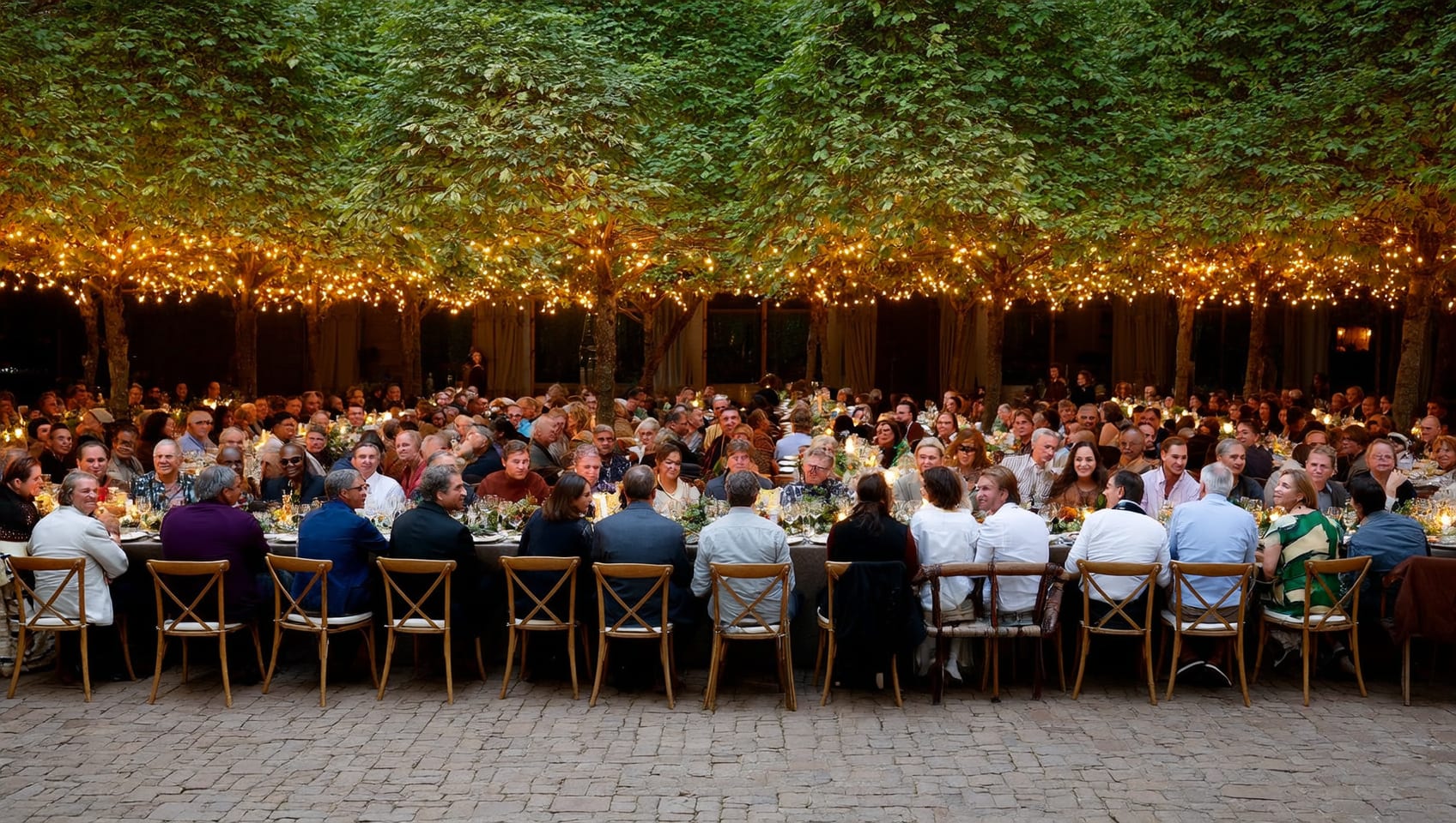A large diverse group seated at a long outdoor dinner table strung with lights beneath the trees